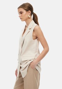 Harmaa hihaton bleiseri, jossa on läppäreunat, etutaskut ja nappikoristeet, yhdistettynä vaaleanruskeisiin housuihin, esitellen sileää kankaan tekstuuria.