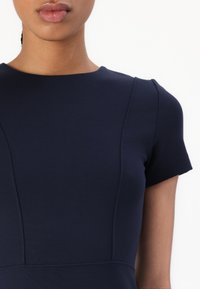 Navy sininen istuva mekko, jossa on lyhyet hihat, pyöreä kaula-aukko ja hienovaraista sauman yksityiskohtaa bodice-osassa, joka luo rakenteellista ilmettä.