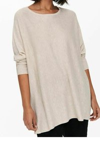 Femme portant un pull ample à manches longues de couleur beige clair avec des poignets côtelés et un pantalon noir, debout les bras détendus le long du corps.
