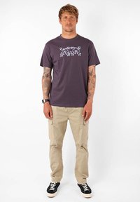 Camiseta de algodón morada con un estampado gráfico, combinada con pantalones cargo beiges que tienen bolsillos laterales y zapatillas negras con suelas blancas.