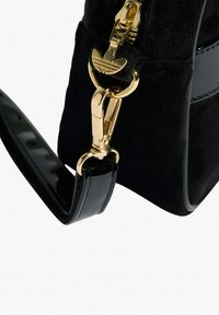 Čierna semišová kabelka s lesklým čiernym remienkom a zlatými kovovými doplnkami. Má výrazný zips na zatváranie s logom. Hladká textúra.