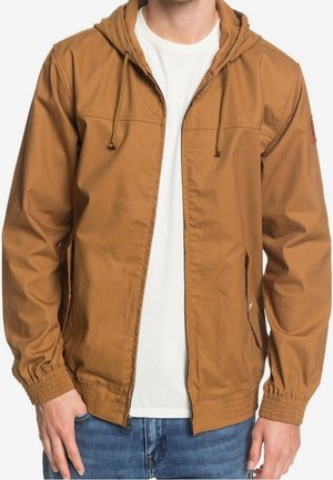 Brun huva jacka i glatt tyg, med framdragkedja, elastiska ärmslut och sidofickor. Bärs över en vit skjorta.