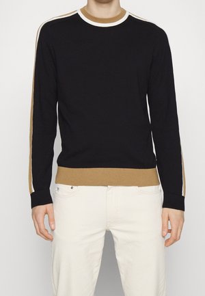 Homme portant un pull noir à manches longues avec des rayures marron et blanches aux manches, au col et à l'ourlet, assorti à un pantalon blanc.