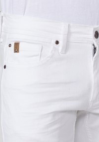 Jeans de denim blanco que presentan un diseño clásico de cinco bolsillos, una discreta etiqueta de marca marrón y herrajes de metal con cremallera. Textura suave.