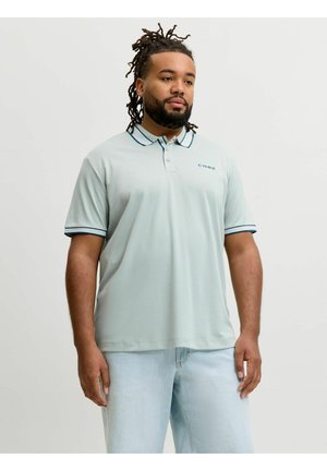 Homme avec des dreadlocks portant un polo bleu clair avec le texte "CORE" et un jean bleu clair, se tenant devant un fond uni.