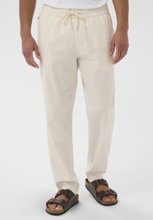 Mężczyzna w beżowych spodniach z wiązaniem i brązowych sandałach z podwójną klamrą, stojący na tle białego, jednolitego tła.