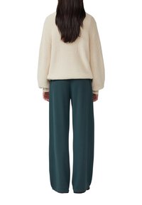 Femme aux longs cheveux noirs portant un pull en tricot crème et un pantalon large vert foncé, se tenant de dos sur un fond blanc.