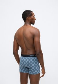 Cuecas boxer azuis com padrão e cós preto com a palavra "HUGO". O design inclui texto repetido em padrão de grade.