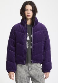 Paarse corduroy pufferjack met een opstaande kraag, gewatteerd ontwerp, elastische manchetten en taille, gedragen over een donker graphic sweatshirt.