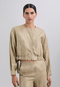 Chaqueta beige corta con mangas largas, dobladillo elástico y adornos plateados decorativos en patrones ondulados. Textura suave, cierre de botón.
