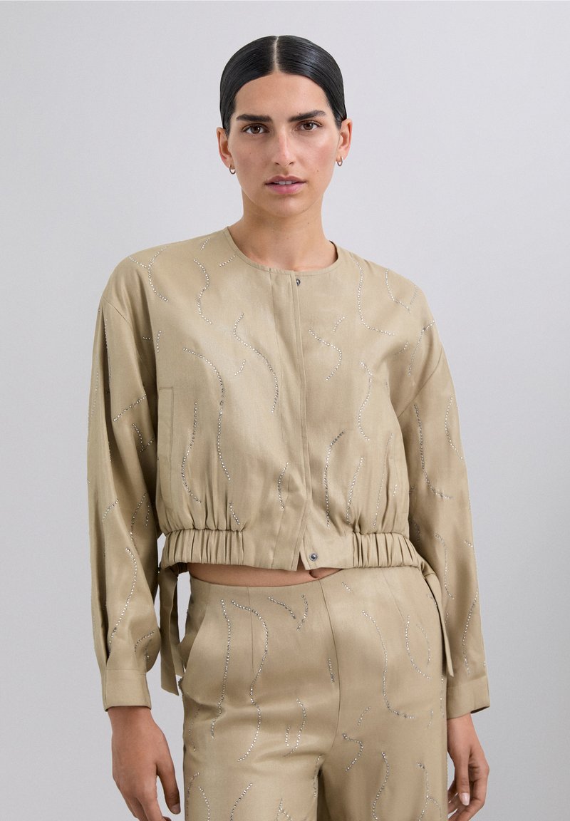 Chaqueta beige corta con mangas largas, dobladillo elástico y adornos plateados decorativos en patrones ondulados. Textura suave, cierre de botón.