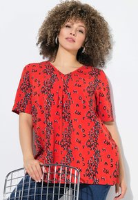 Top corto rojo de manga corta con escote en V, con patrones de texto negro en toda la prenda. Tejido suave con caída en la parte delantera, combinado con Jeans azules.