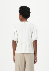 Weiße strukturierte Bluse mit Puffärmeln, Peplum-Saum und einem kleinen Rückenetikett. Kombiniert mit hellbeigen Hosen.