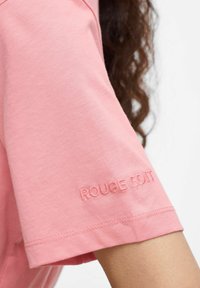 T-shirt corto a maniche corte di colore rosa chiaro, realizzato in cotone, con testo ricamato "ROUGE EDIT" sulla manica. Tessuto morbido, vestibilità casual.