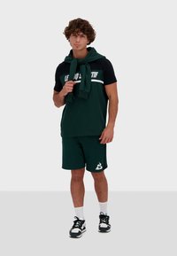 Conjunto deportivo en verde y negro que incluye una camiseta de manga corta con capucha y pantalones cortos a juego, con un detalle de logo y zapatillas casuales.