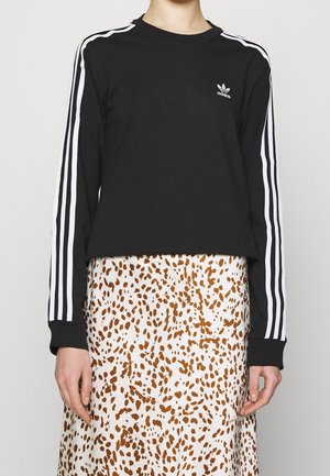 Person trägt ein schwarzes Adidas-Langarmshirt mit weißen Streifen an den Ärmeln und einen weißen Rock mit braunem Fleckenmuster.