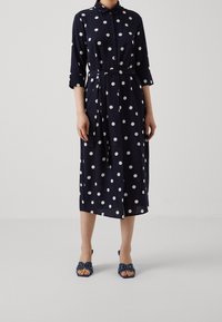 Donna che indossa un abito midi blu navy con pois bianchi, maniche a tre quarti, un nastro in vita e sandali con tacco aperti coordinati.