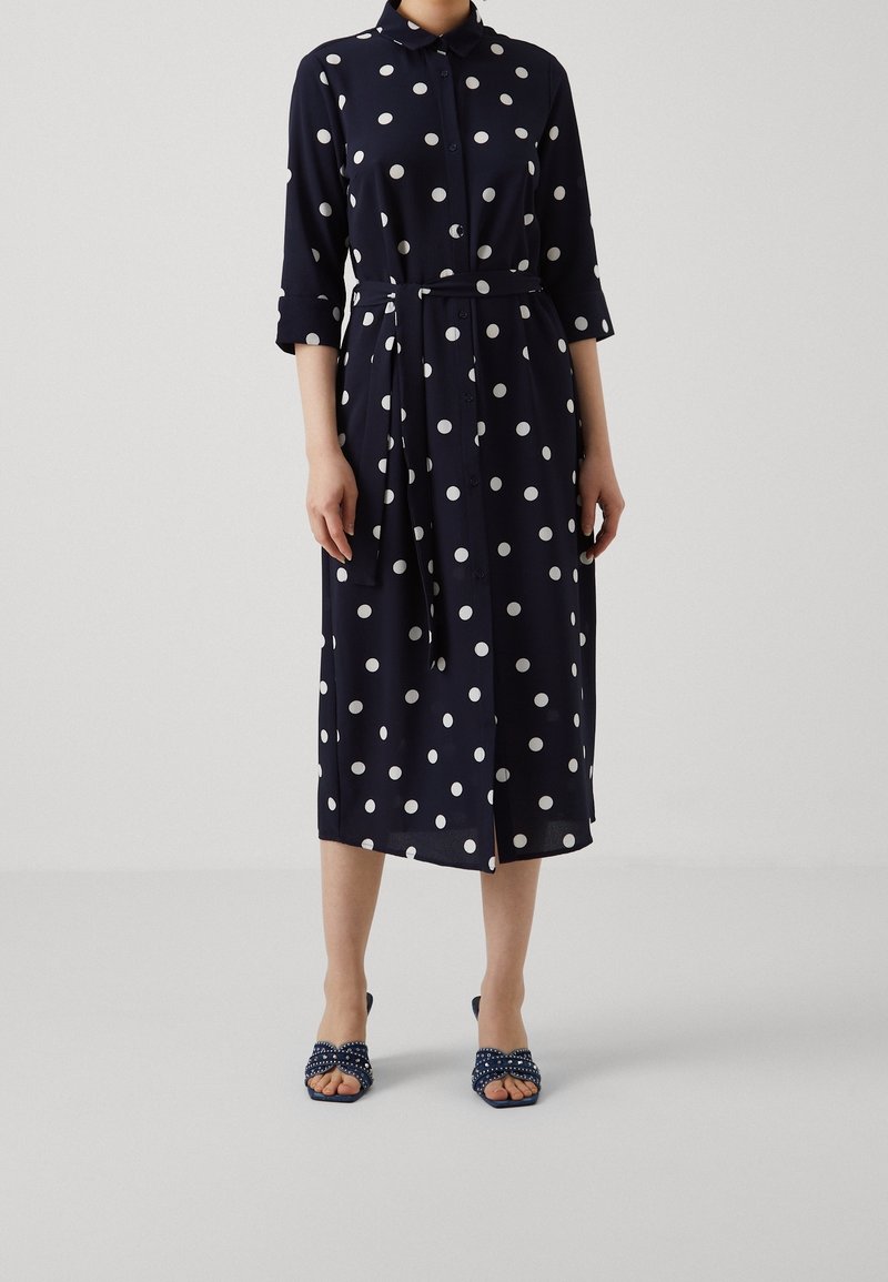 Donna che indossa un abito midi blu navy con pois bianchi, maniche a tre quarti, un nastro in vita e sandali con tacco aperti coordinati.