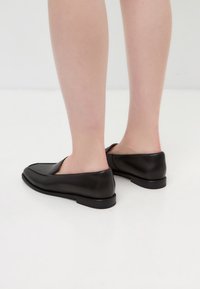 Black leather slip-on shoes with a smooth texture, square toe design, and minimalistic low profile, viewed from the back.