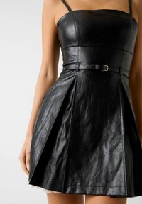 Robe en cuir noir avec fines bretelles, corsage ajusté, jupe plissée et taille ceinturée avec un détail subtil de boucle métallique.