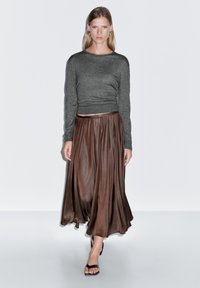 Šedý top s dlouhým rukávem spojený s hnědou saténovou sukní, která lehce splývá. Sukně má jemný lesk a sahá pod kotníky, kde odhaluje černé sandály.
