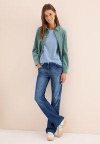 Veste boutonnée en velours côtelé vert sur un t-shirt bleu clair, associée à un jean évasé bleu et des baskets beiges avec des accents bleus.
