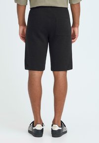 Shorts noirs en tissu texturé, avec une poche arrière. Portés avec des baskets noires et blanches, affichant un design simple et fonctionnel.