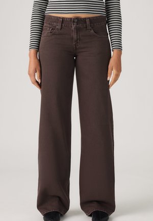 Femme portant un jean marron foncé taille haute à jambes larges avec une fermeture à double bouton et un haut à manches longues rayé noir et blanc.