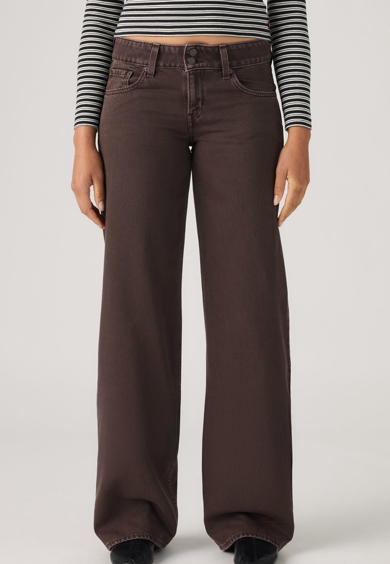Femme portant un jean marron foncé taille haute à jambes larges avec une fermeture à double bouton et un haut à manches longues rayé noir et blanc.