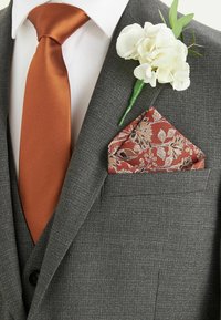 Un costume gris texturé avec une cravate en satin orange, un mouchoir de poche floral avec des motifs rouges et dorés, et une boutonnière en fleur blanche.