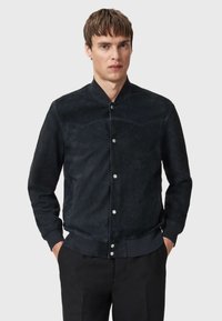Giubbotto bomber in suede scuro con colletto alto, bottoni a pressione e polsini a coste. Caratterizzato da una texture liscia e linee pulite.