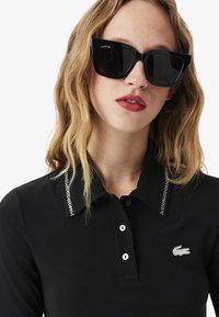 Zwarte poloshirt met een kraag versierd met zilveren embellishments, voorzien van een verborgen knoopsluiting en een geborduurd Lacoste-logo op de borst.