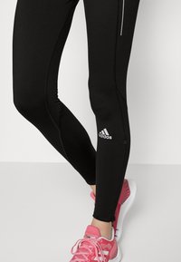 Czarne legginsy sportowe o dopasowanym kroju, z odblaskowymi akcentami i logo Adidas. W zestawieniu z różowymi, teksturowanymi butami do biegania.