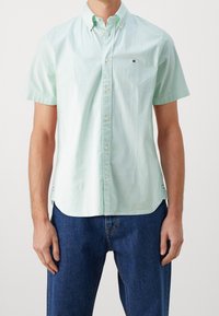 Camisa de botones de manga corta en verde claro con franjas blancas delgadas alternas. Presenta un cuello y un pequeño logo en el pecho.