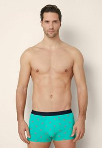 Boxer da uomo verdi con motivo a cocomero, elastico blu scuro e tessuto morbido. Il modello è in piedi con un'espressione neutra e il torsocn nudo.