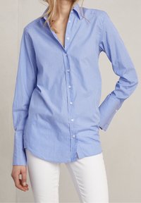 Světle modrá košile na knoflíky s bílými svislými pruhy. Obsahuje špičatý límec, dlouhé rukávy a zaoblený lem s uvolněným střihem.