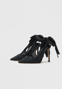 Pantofi cu toc înalt negri, cu vârf ascuțit, decorați cu paiete, având o fundă cu șireturi în spate și un toc stiletto elegant.