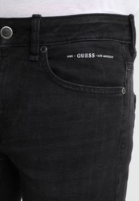 Jeans de denim negro con un corte ajustado, diseño de cinco bolsillos y un logo blanco "GUESS" en la cinturilla. Cierre con botón de latón y cremallera.
