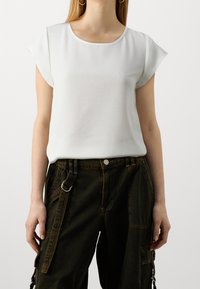 Top corto bianco a maniche corte con scollatura rotonda e texture liscia, abbinato a pantaloni cargo verde scuro con diverse tasche e dettagli in metallo.