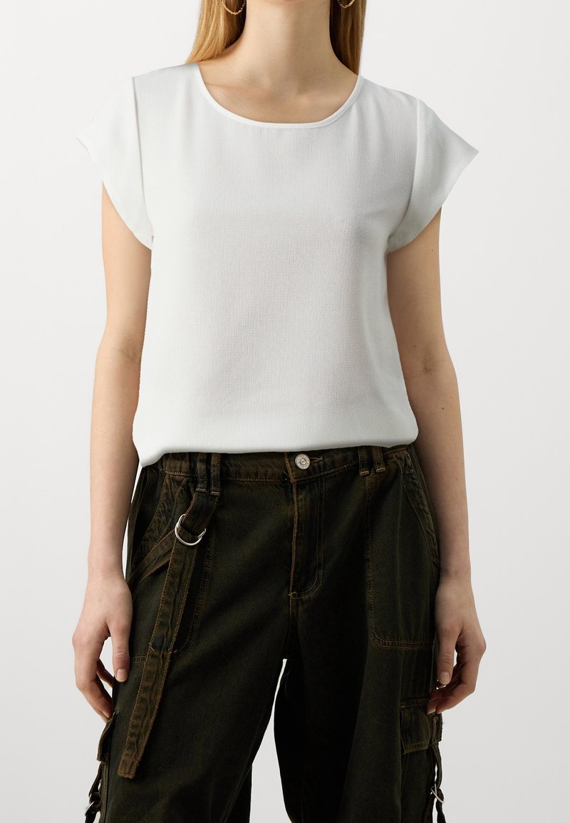 Top corto bianco a maniche corte con scollatura rotonda e texture liscia, abbinato a pantaloni cargo verde scuro con diverse tasche e dettagli in metallo.