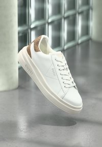 Baskets basses blanches avec talon texturé marron, lacets blancs et logo "GUESS" embossé sur le côté et la semelle, posées sur un sol réfléchissant.