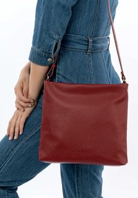 Sac à bandoulière en cuir rouge avec une finition texturée, de forme rectangulaire et une longue sangle. Positionné contre une silhouette vêtue de denim.