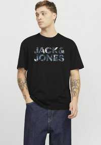Mustat puuvillaiset t-paidat, joissa on "JACK & JONES" kuvioitu painatus. Lyhyet hihat ja pyöreä kaula-aukko, näkyvissä siniset farkut.