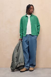 Grøn struktureret skjorte med knapper over en hvid t-shirt, kombineret med løse blå denimjeans og beige sko. Grå bomberjakke holdt i hånden.