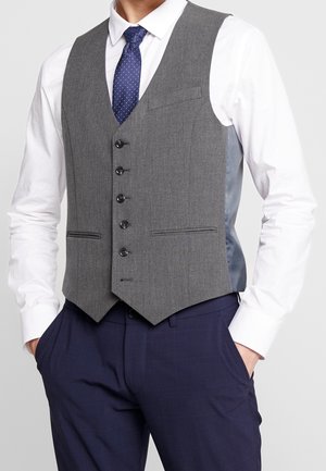 Mężczyzna w szarej kamizelce na guziki, białej koszuli, granatowym krawacie w groszki i granatowych eleganckich spodniach, z rękami w kieszeniach.