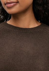 Primo piano della parte inferiore del volto e del collo di una persona che indossa un maglione di lana marrone, con capelli scuri e ricci visibili.