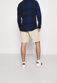 Camicia a quadri nera e blu a maniche lunghe con dettagli a bottoni, abbinata a pantaloncini beige. Scarpe da ginnastica bianche con accenti neri.