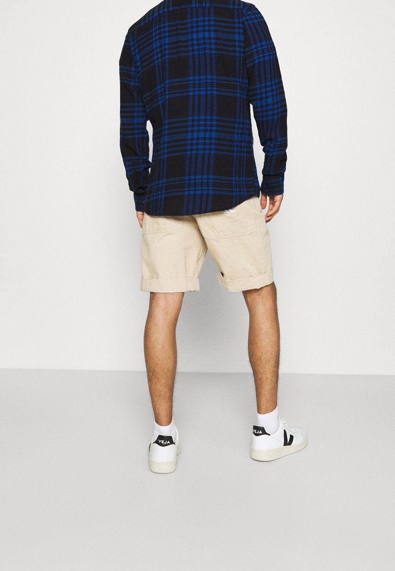 Camicia a quadri nera e blu a maniche lunghe con dettagli a bottoni, abbinata a pantaloncini beige. Scarpe da ginnastica bianche con accenti neri.