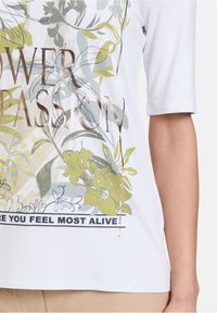 Hvid kortærmet t-shirt med blomstermønster i grøn og beige, med metallisk guldtekst der siger "FLOWER PASSION" og en sort accent nedenfor.