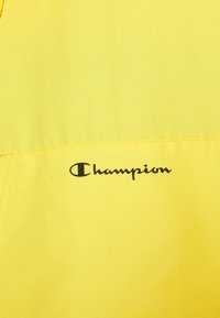 Klart gult tyg med en slät yta har en svart "Champion"-logotyp. Designen är enkel med minimala sömnadsdetaljer.